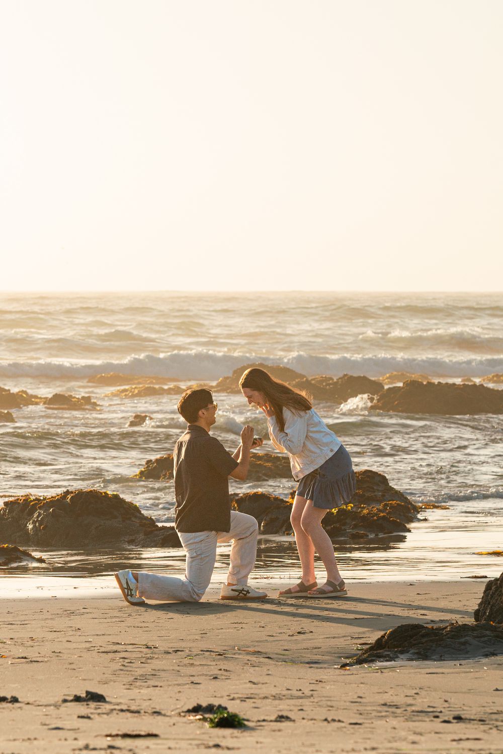 Man proposes to woman on one knee at a golden sunset beach, waves crashing on rocks behind them.