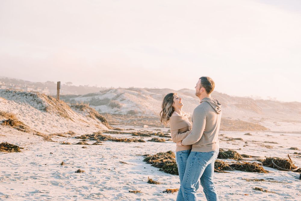 Couple embracing on a sandy beach at golden hour, sharing a romantic moment with soft warm light and coastal dunes behind them.