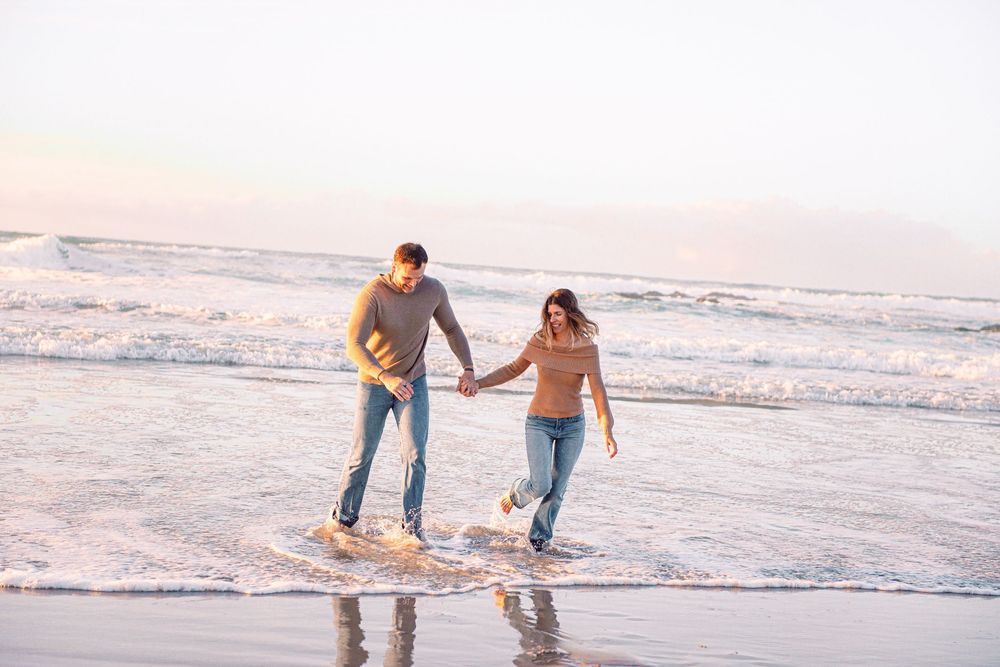 Couple holding hands and walking along a sandy beach shore with ocean waves in the background.