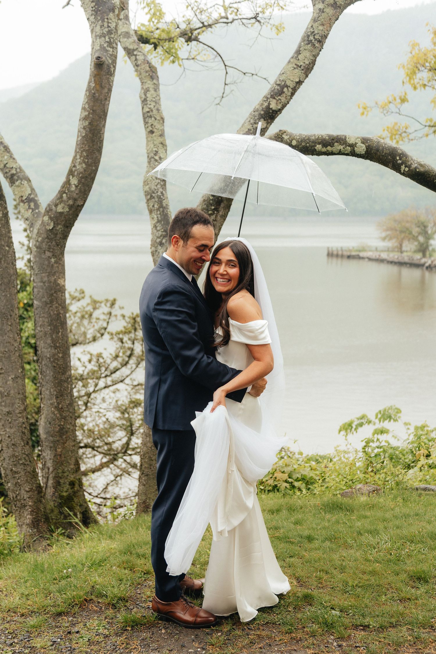 Bride and groom sharing a romantic moment under a clear umbrella by a misty lake on their rainy wedding day.