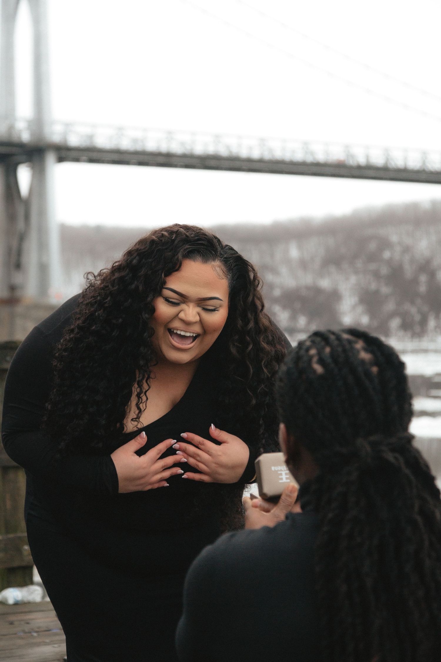 A joyful woman laughs during a marriage proposal near a bridge, with her partner holding an open ring box.