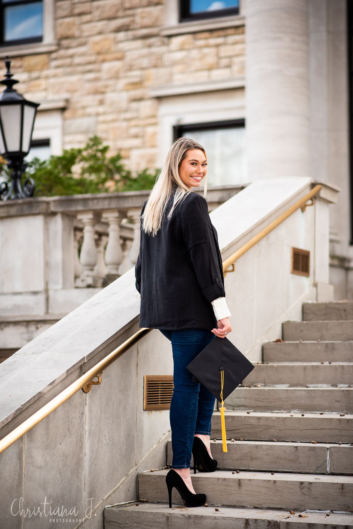 Blonde graduate in black blazer holding cap while descending elegant stone staircase.