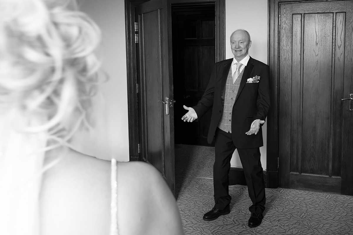 Black and white photo of an older man in a suit smiling at a bride during a wedding day first look moment.