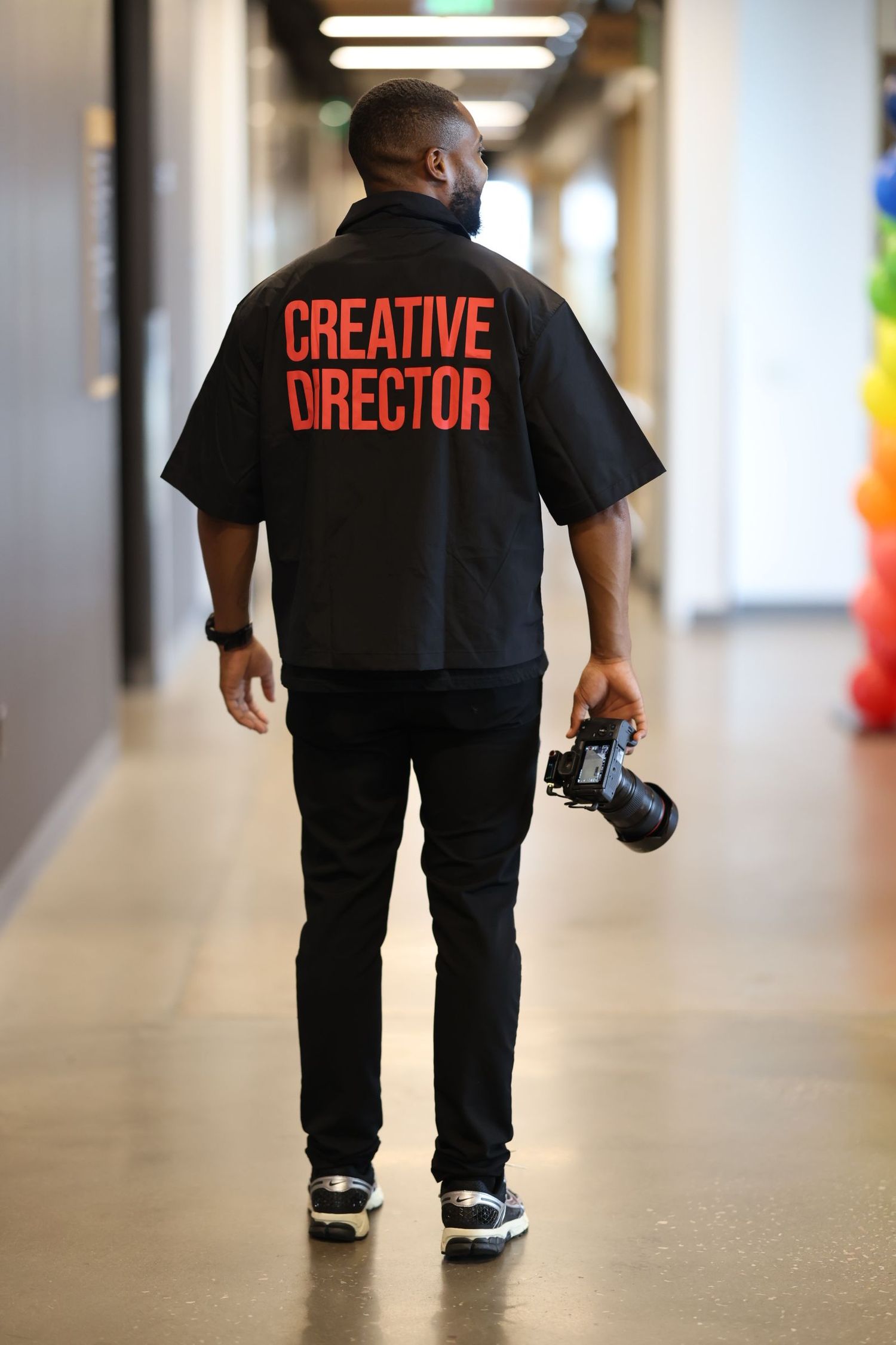 Man wearing black Creative Director shirt holding camera walks down a hallway.