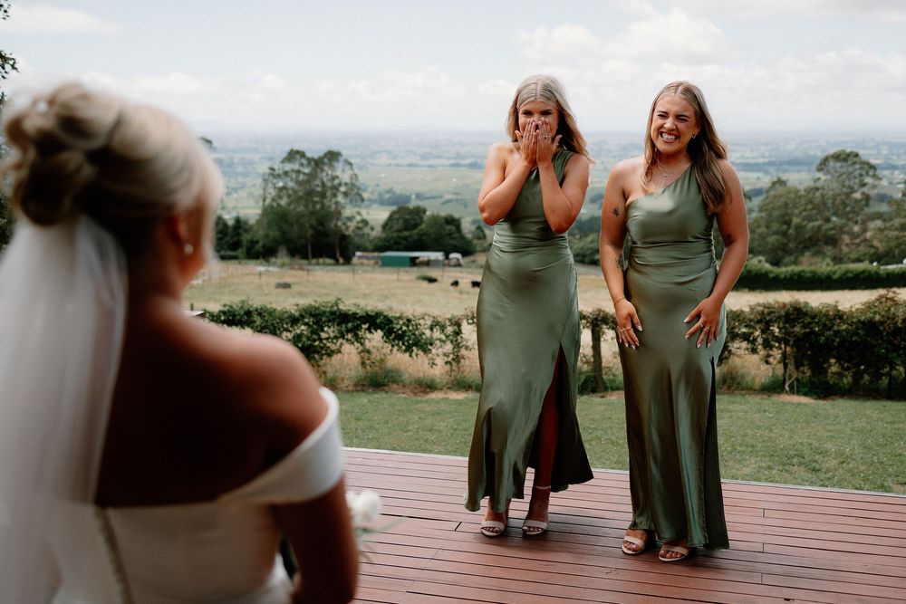 Bridesmaids in matching sage green dresses react with joy on a wooden deck overlooking a scenic countryside view.