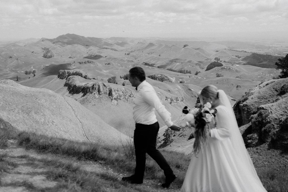 A couple in wedding attire walks along a scenic mountain ridge with rolling hills stretching into the distance.