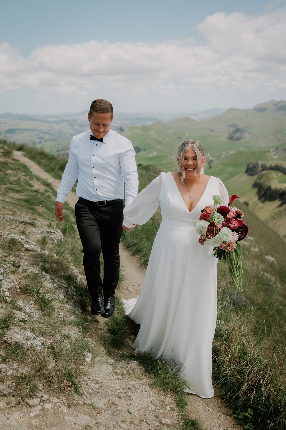 Wedding couple walking along scenic mountain ridge trail with rolling green hills in background.