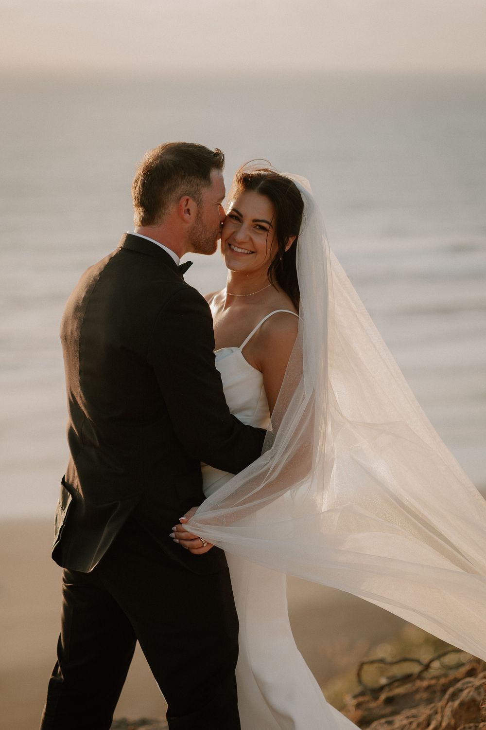 A romantic wedding couple embraces on a beach at sunset as the bride's veil flows dramatically in the wind.