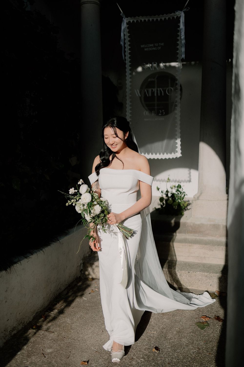 A bride in a white wedding dress walks gracefully down a shadowy corridor holding a delicate bouquet of white flowers.