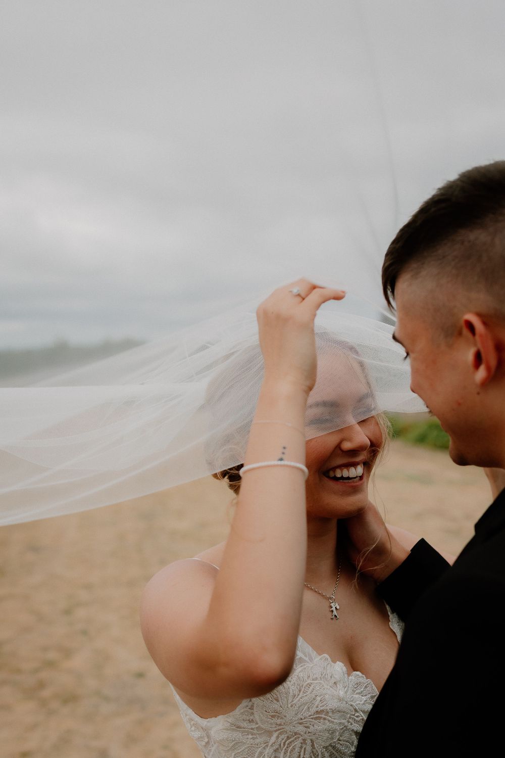 A joyful wedding couple shares an intimate moment on a cloudy beach as the bride's veil flows in the wind.