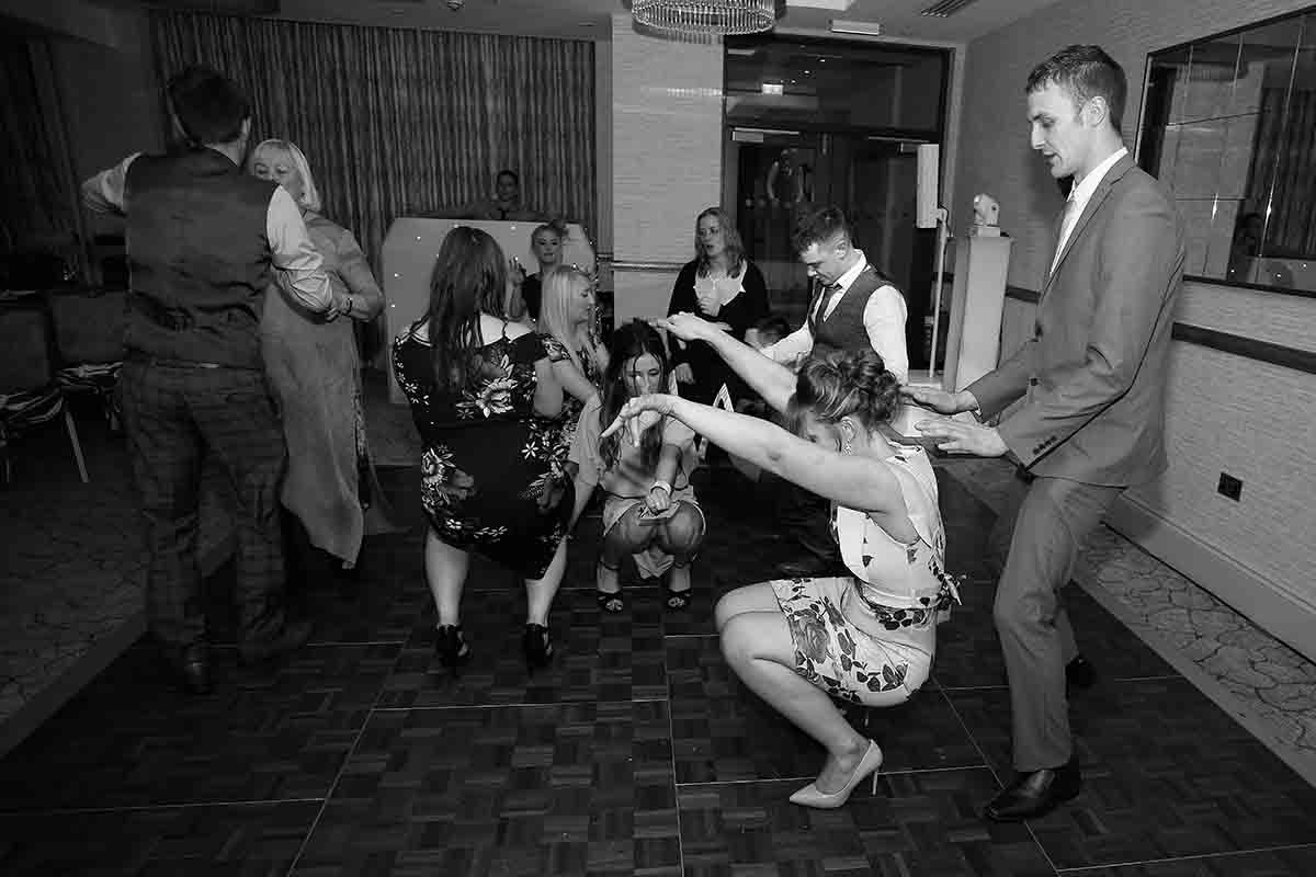 Black and white photo of guests dancing energetically at a lively wedding reception party.