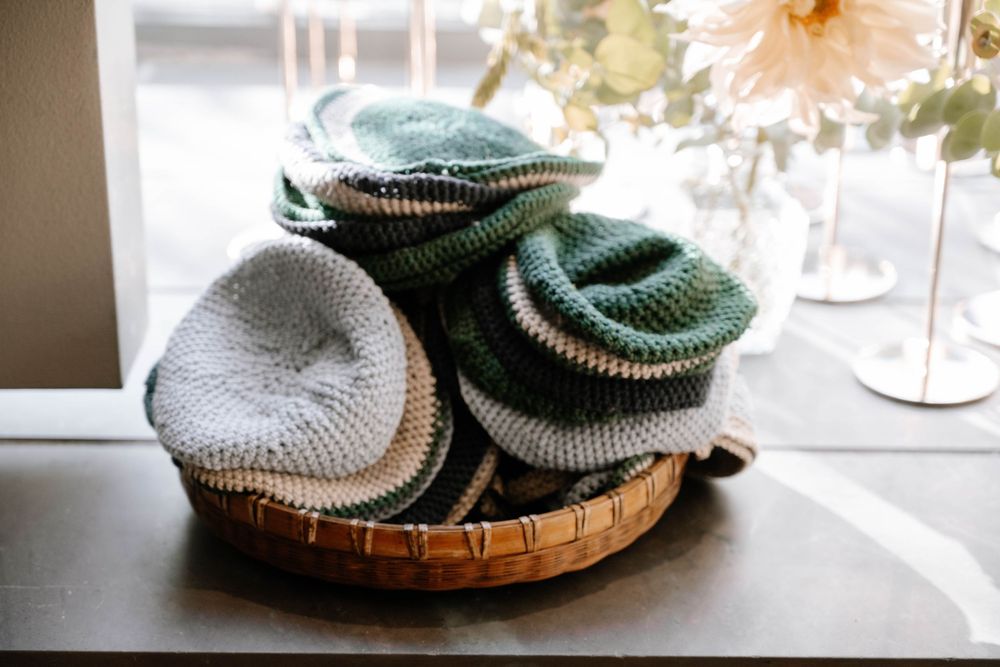 Handmade crochet beanies in green and gray tones displayed in a woven basket on a windowsill.