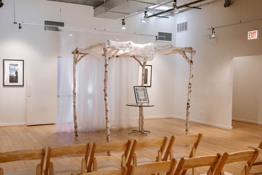 Elegant Jewish wedding chuppah with birch poles and white fabric draping in a gallery space with wooden chairs.