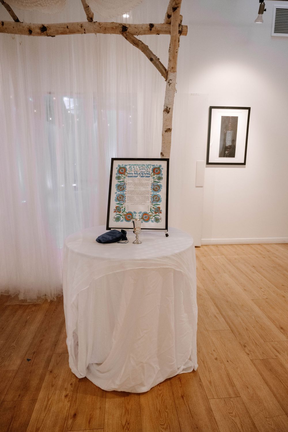 A white-draped round table with a framed floral document and birch tree branches in an elegant gallery space.