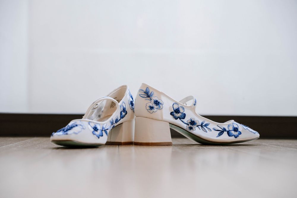 White heeled shoes with blue floral embroidery displayed on a wooden floor.
