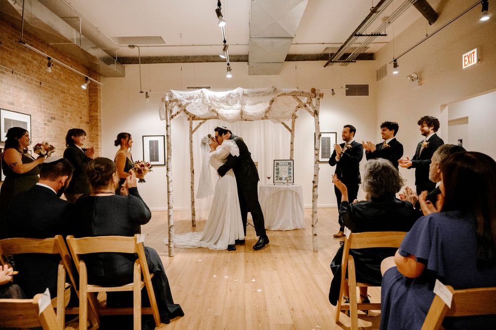 Bride and groom share first kiss under floral chuppah at elegant indoor Jewish wedding ceremony with guests applauding.