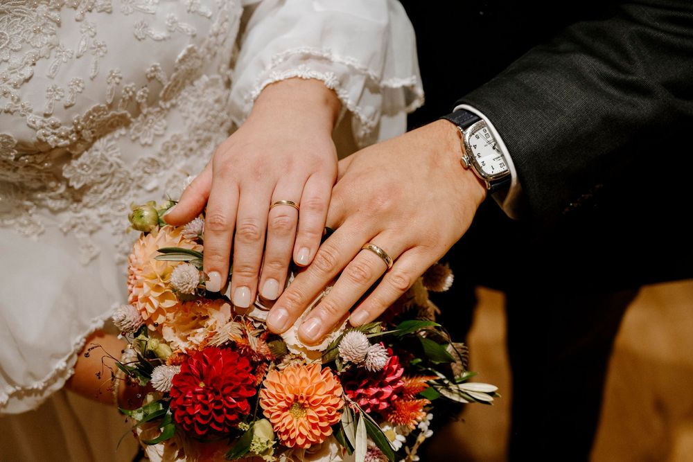 Newlywed couple displaying gold wedding rings on hands resting over a colorful bridal bouquet with red dahlias.