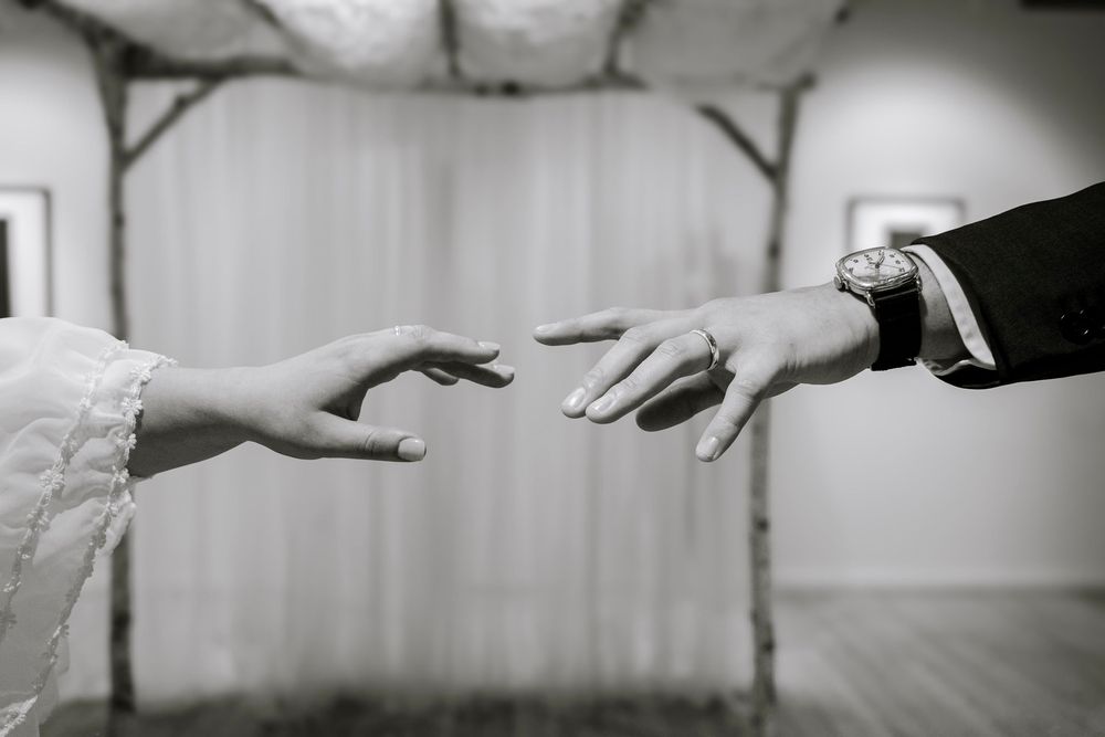 Black and white photo of bride and groom reaching hands toward each other, groom wearing a watch and wedding ring.