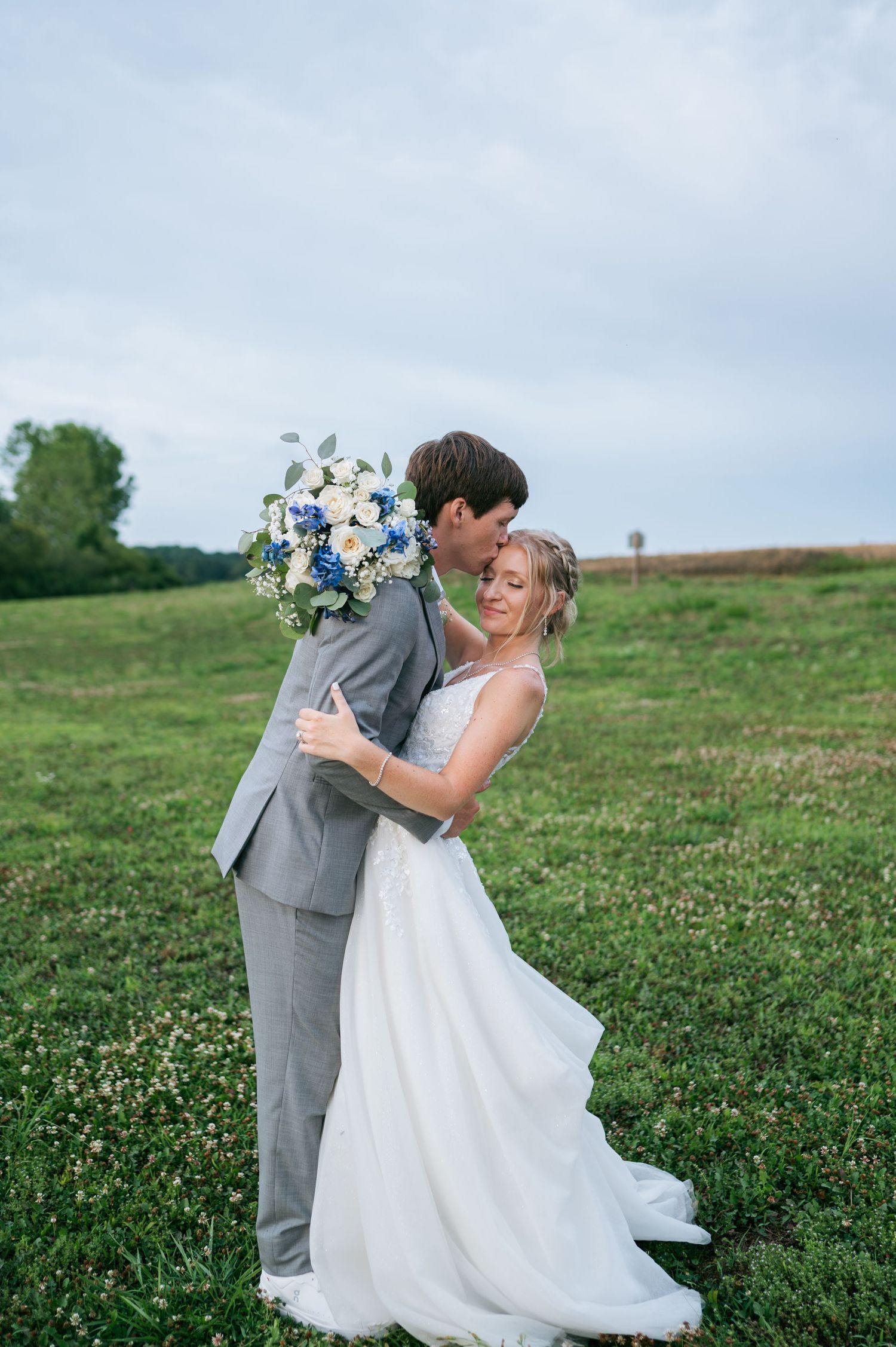 Groom in gray suit kisses bride in white gown while she smiles holding a blue and white bouquet in a green field, wedding photographer NC