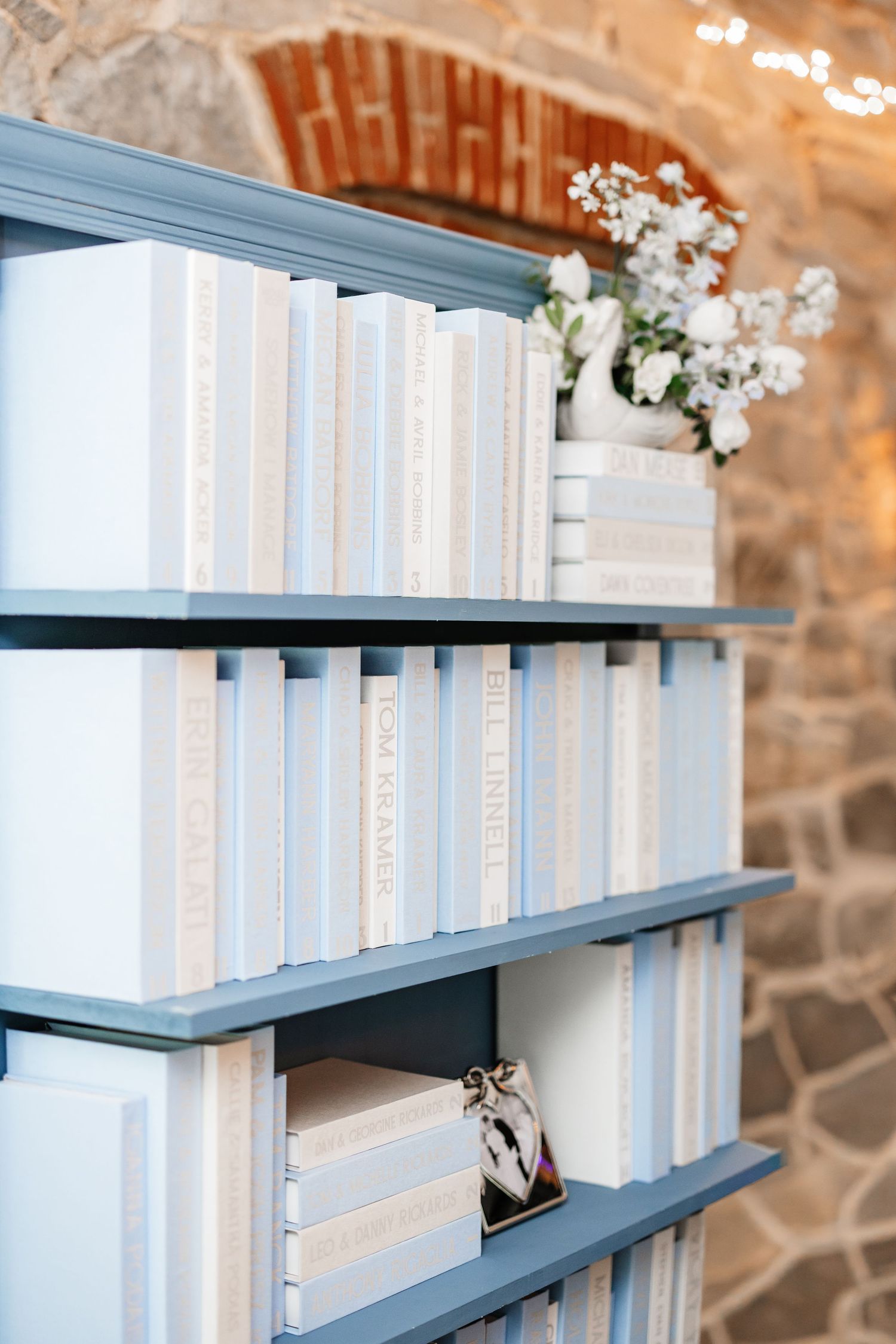 Blue bookshelf displaying white and cream books with small white floral arrangement as wedding decor.