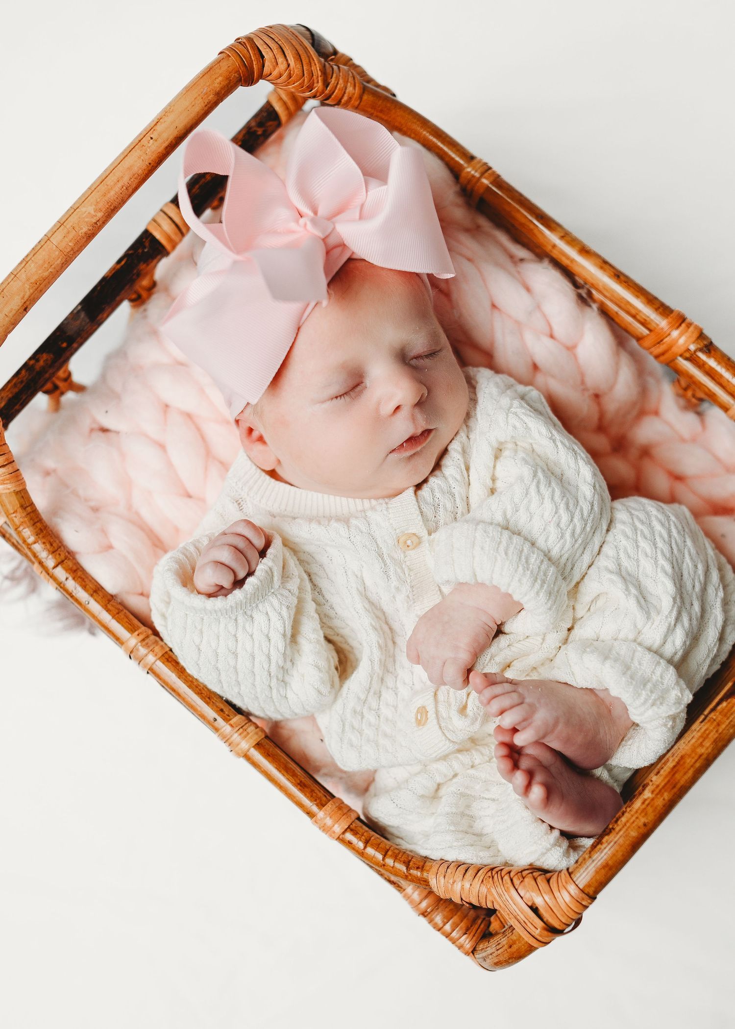 Sleeping newborn baby girl in white knit outfit and pink bow headband resting in a wicker basket.