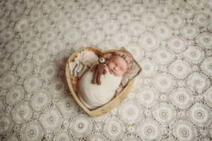 Newborn baby swaddled in white wrap sleeping in heart-shaped wooden bowl with teddy bear on lace backdrop.