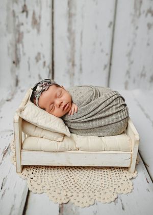 Sleeping newborn baby swaddled in gray wrap with floral headband resting on tiny white wooden bed with cream pillow.