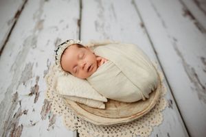 Sleeping newborn baby swaddled in cream wrap, nestled in wooden bowl with lace, wearing delicate bonnet.