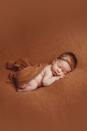 Sleeping newborn baby girl wrapped in a rust brown blanket with a delicate gold headband on a matching brown backdrop.