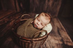 Newborn baby swaddled in green wrap sleeping peacefully inside a wicker basket on rustic wood.