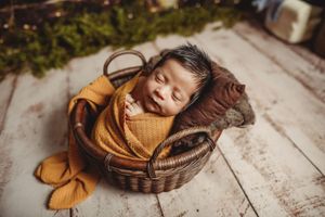 Newborn baby wrapped in mustard yellow swaddle sleeping peacefully in a rustic wicker basket.