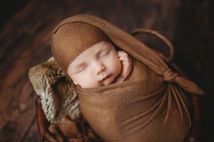Newborn baby sleeping peacefully wrapped in a brown knit swaddle and matching hat inside a rustic wooden bucket.
