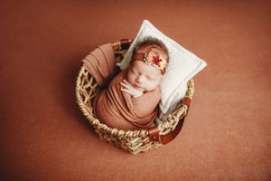 Newborn baby swaddled in rust wrap sleeping in wicker basket with white pillow and floral headband.