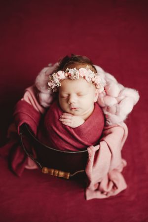 Sleeping newborn baby girl wrapped in pink swaddle wearing floral crown in wooden bowl on red background.