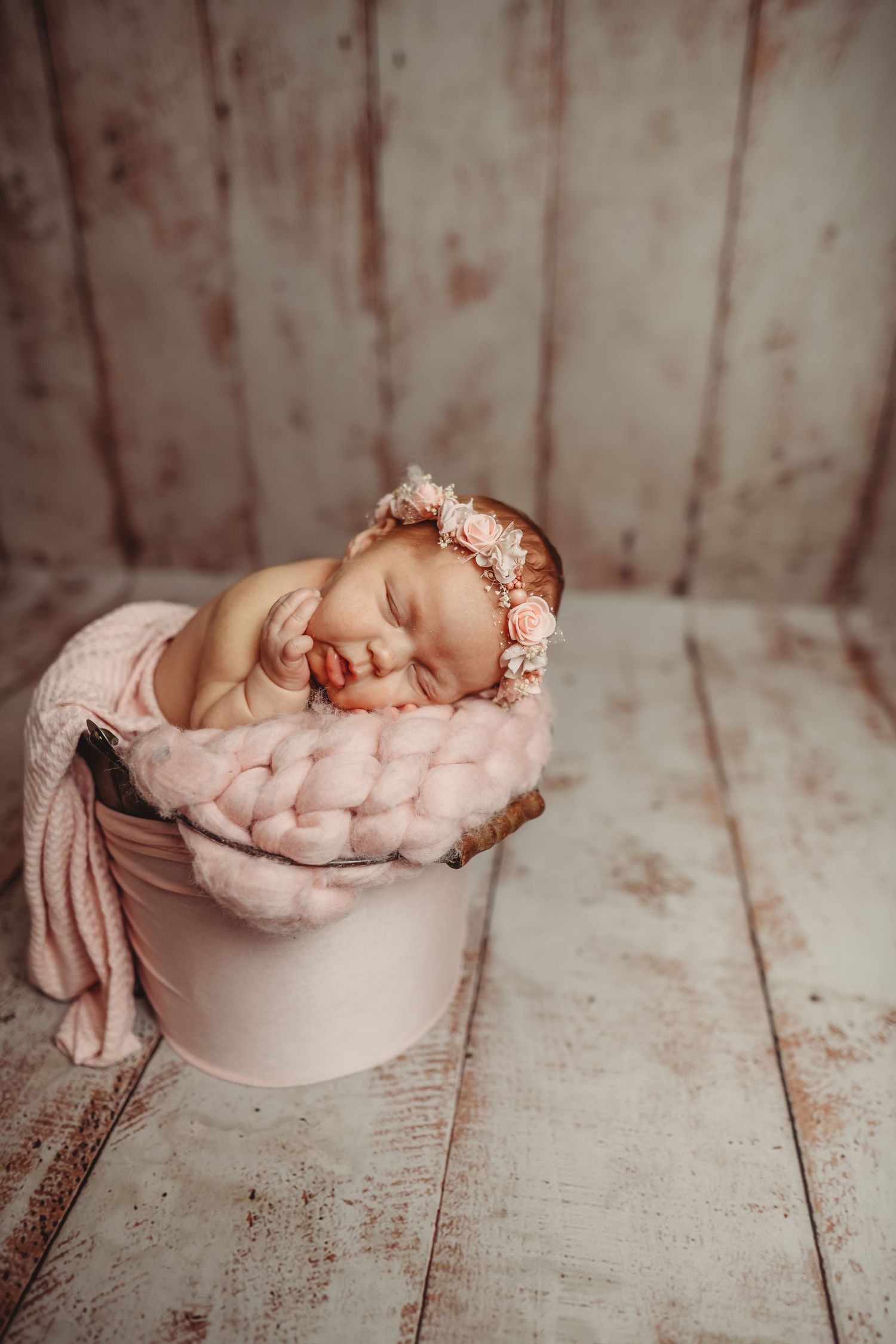 Sleeping newborn baby girl in pink bucket with chunky knit blanket and floral headband on rustic wood background.