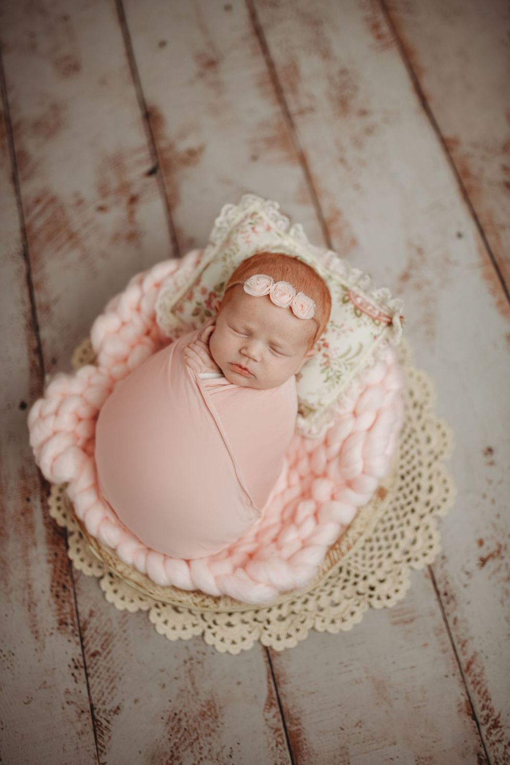 Newborn baby girl swaddled in pink wrap sleeping in a basket with chunky knit blanket and floral bonnet.