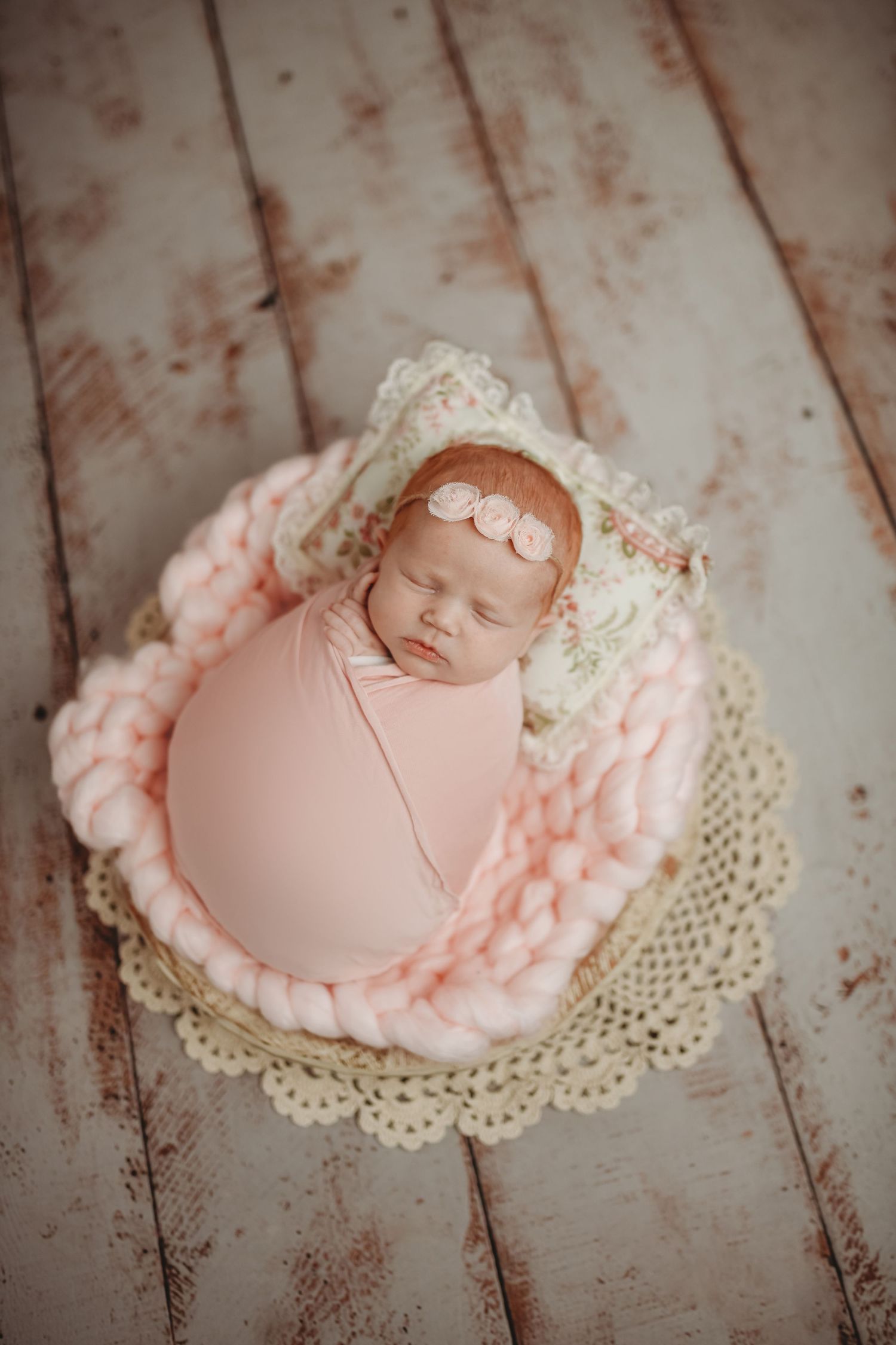 Newborn baby girl swaddled in pink wrap sleeping in a basket with chunky knit blanket and floral bonnet.