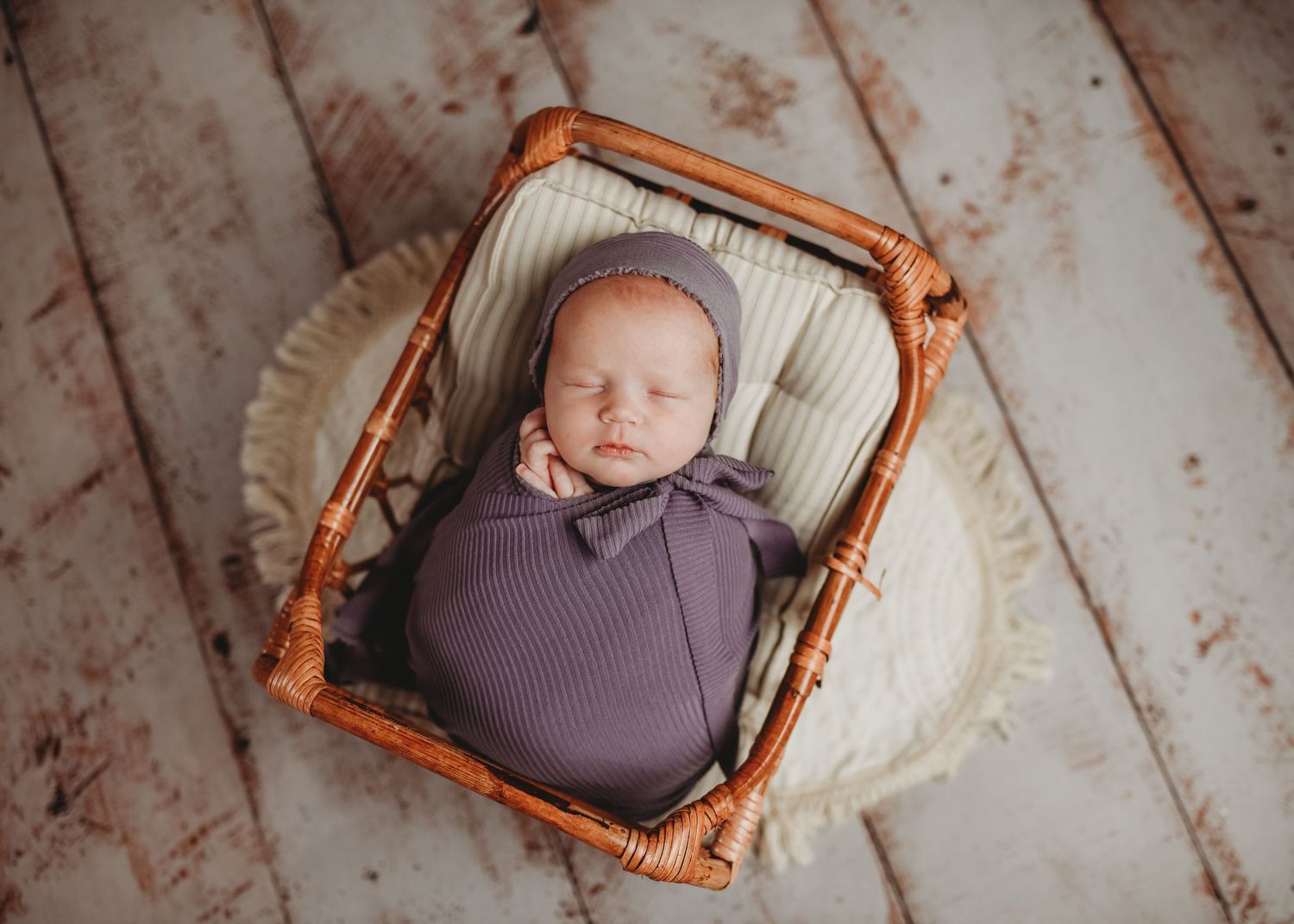 Newborn baby swaddled in purple wrap wearing a bonnet, sleeping peacefully in a rustic wicker basket.