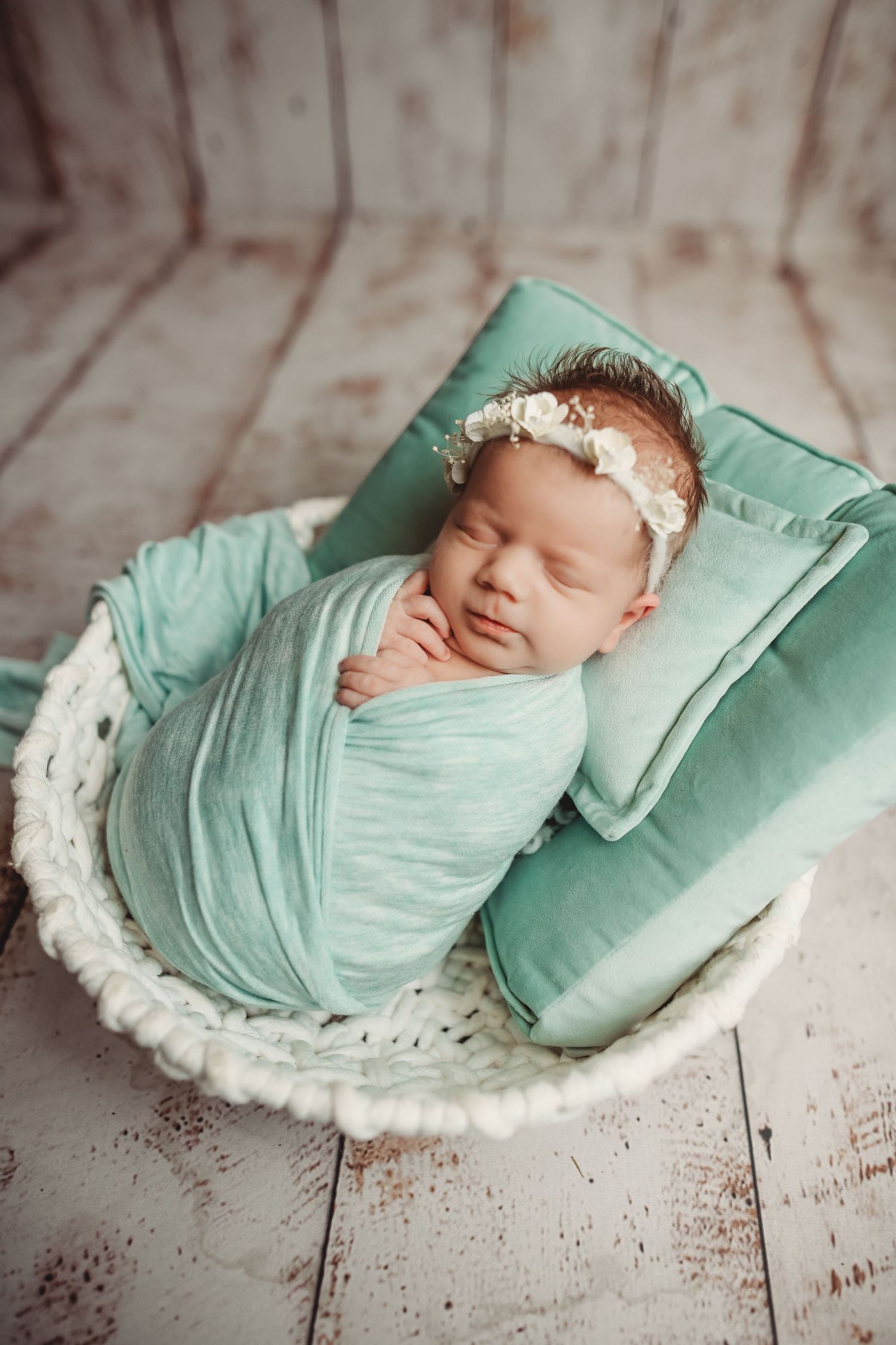 Sleeping newborn baby swaddled in teal wrap with floral headband resting in white wicker basket.