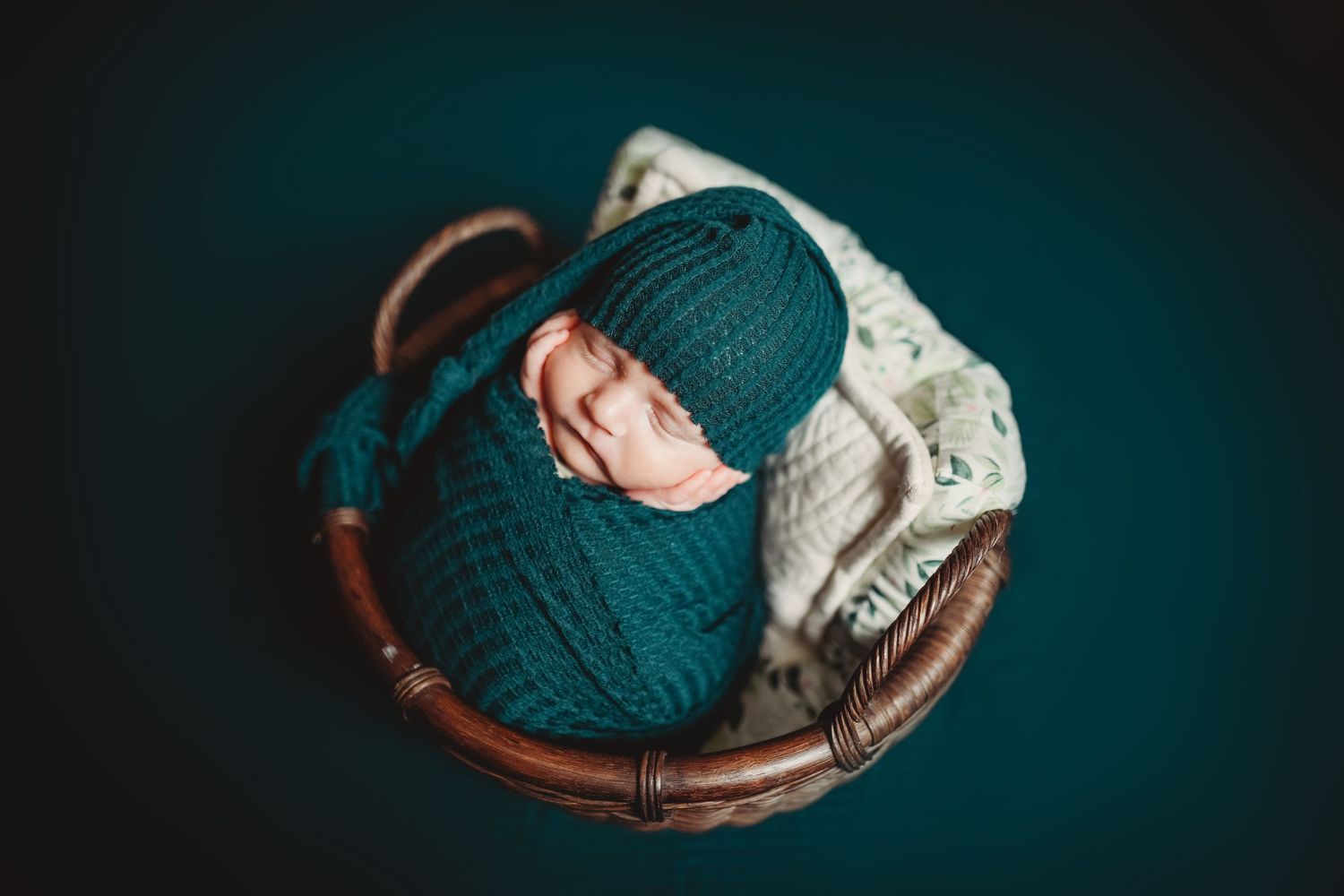 Newborn baby swaddled in teal knit wrap and hat, sleeping peacefully in a wicker basket on dark background.