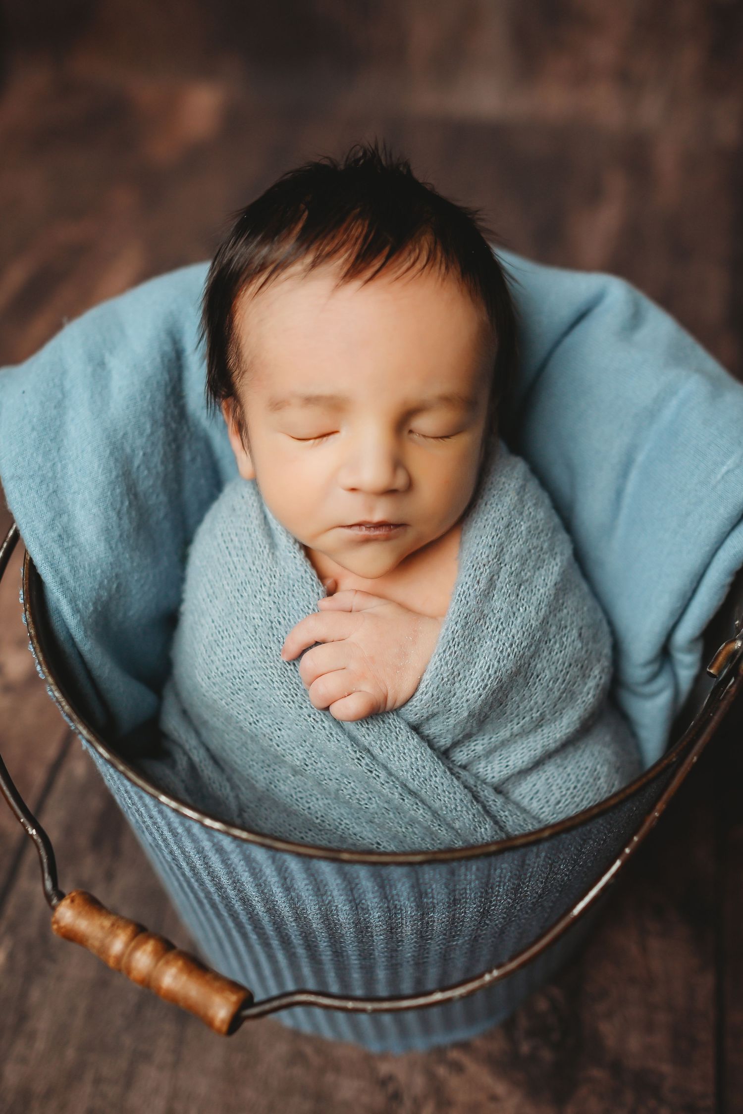 Sleeping newborn baby swaddled in blue wrap inside a rustic metal bucket on wooden floor.