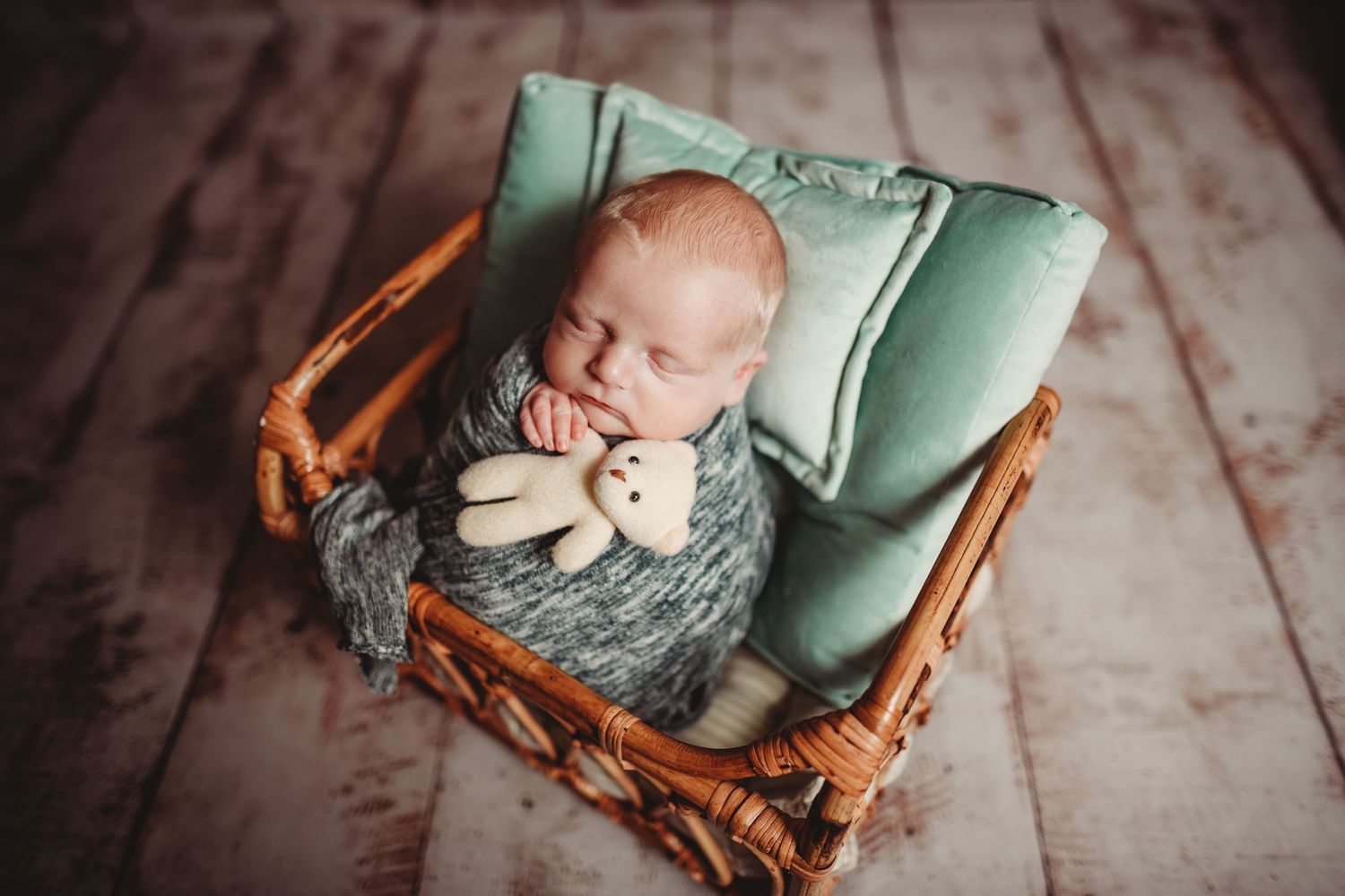 Newborn baby sleeping peacefully in a wicker basket with mint green bedding and a small teddy bear.