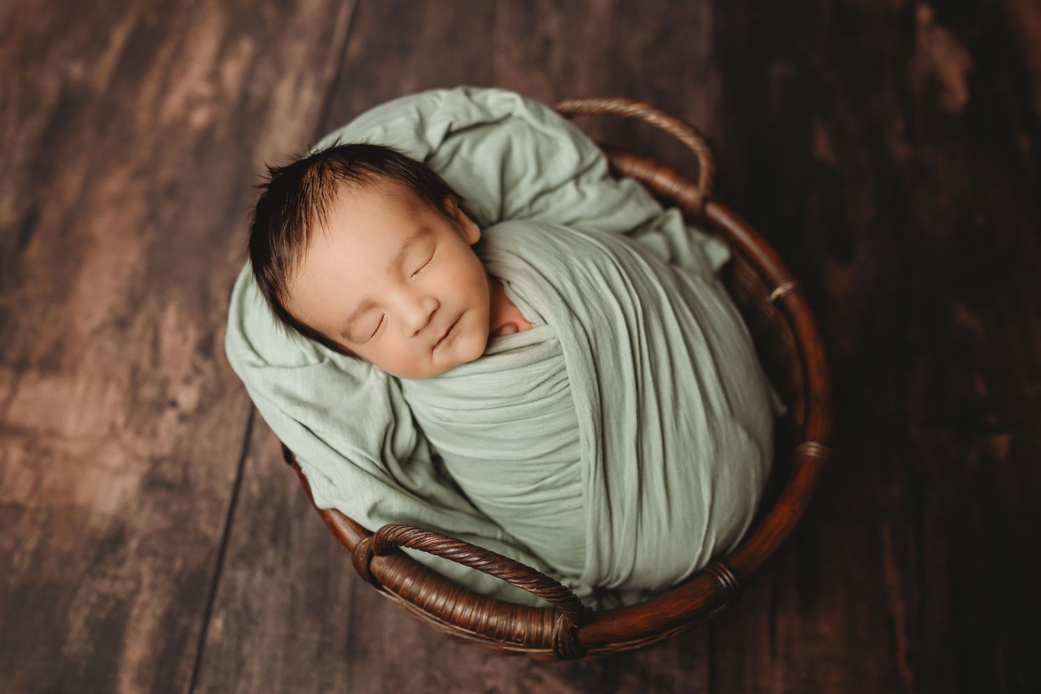 Sleeping newborn baby swaddled in sage green wrap, nestled in a wicker basket on a rustic wood floor.