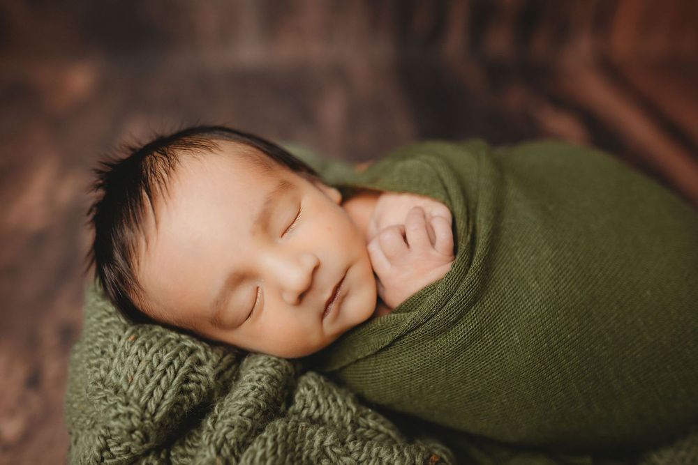 Peaceful newborn baby swaddled in a green knit wrap, sleeping soundly in a cozy wooden bowl.
