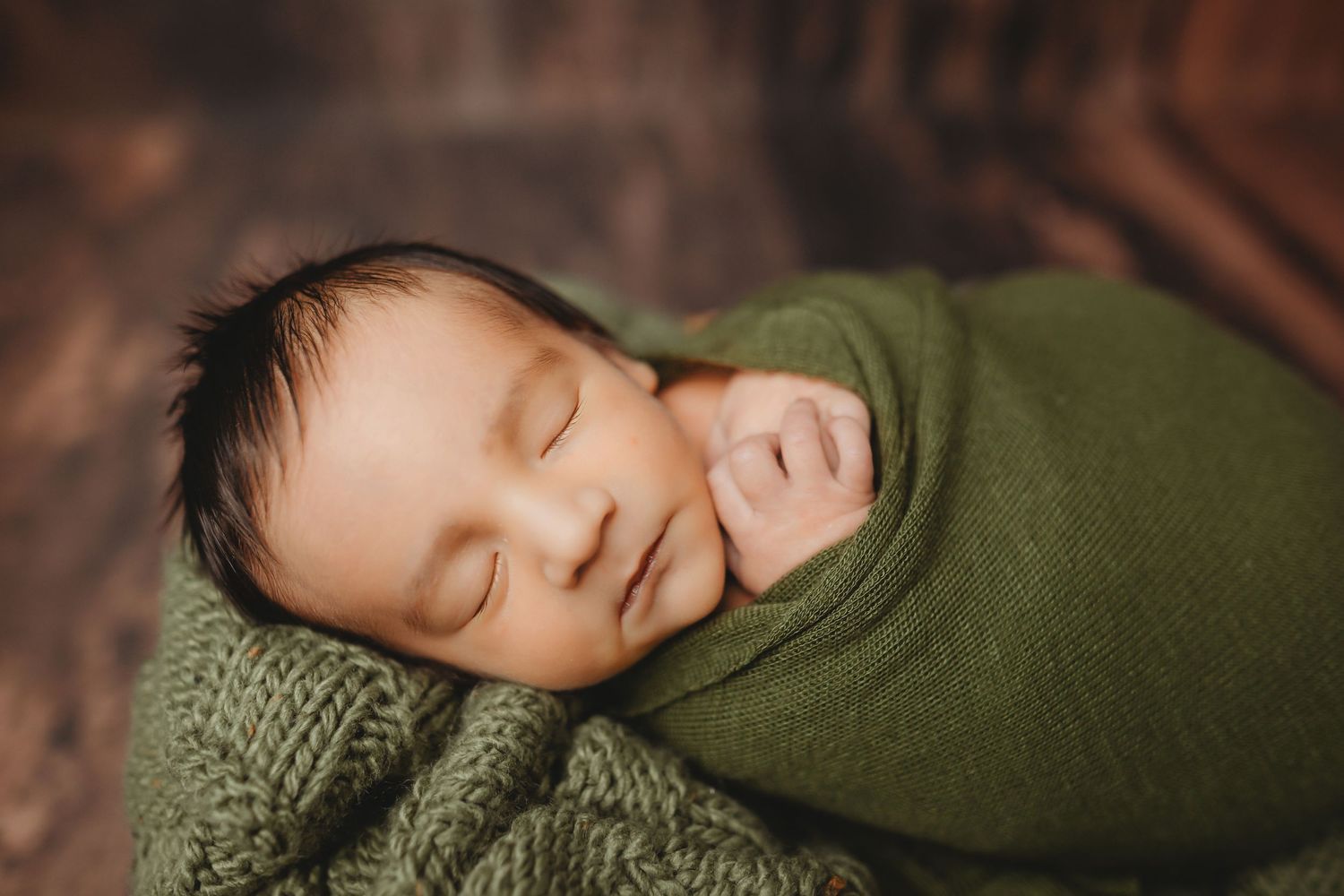 Peaceful newborn baby swaddled in a green knit wrap, sleeping soundly in a cozy wooden bowl.