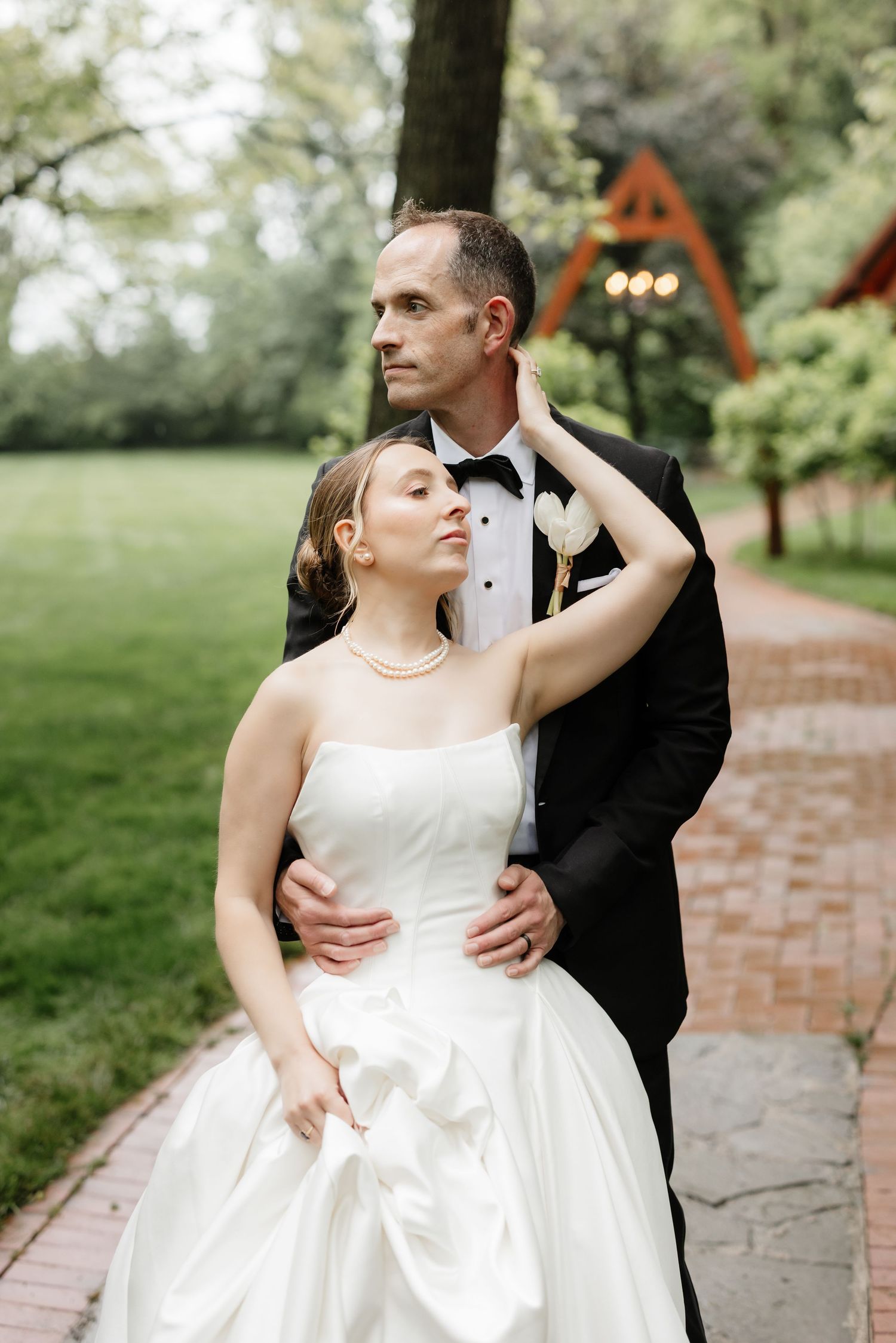 Elegant bride and groom embracing on garden path with wooden arch and greenery backdrop.
