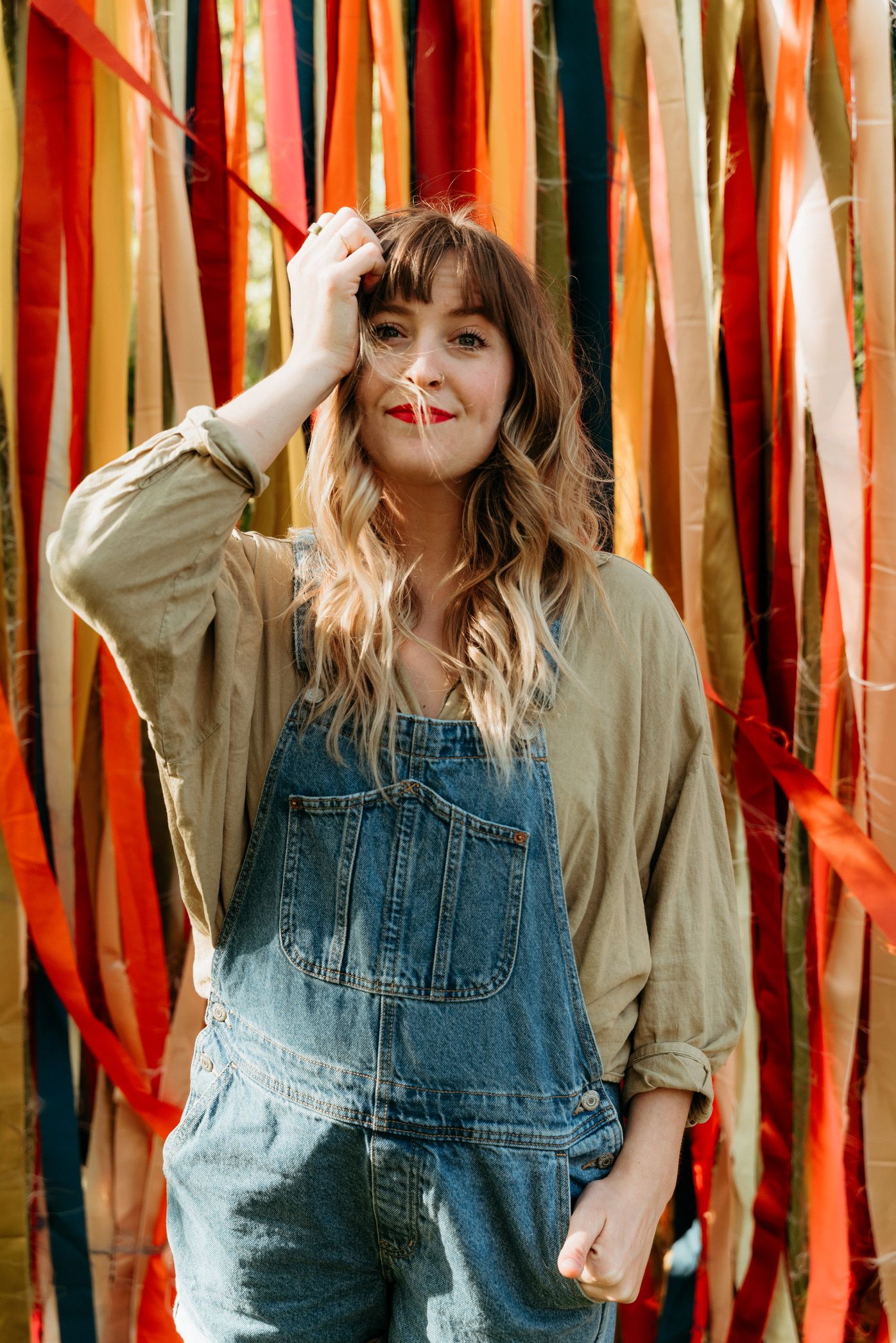 Woman in denim overalls and olive shirt poses before colorful ribbon backdrop with wavy hair and red lipstick.