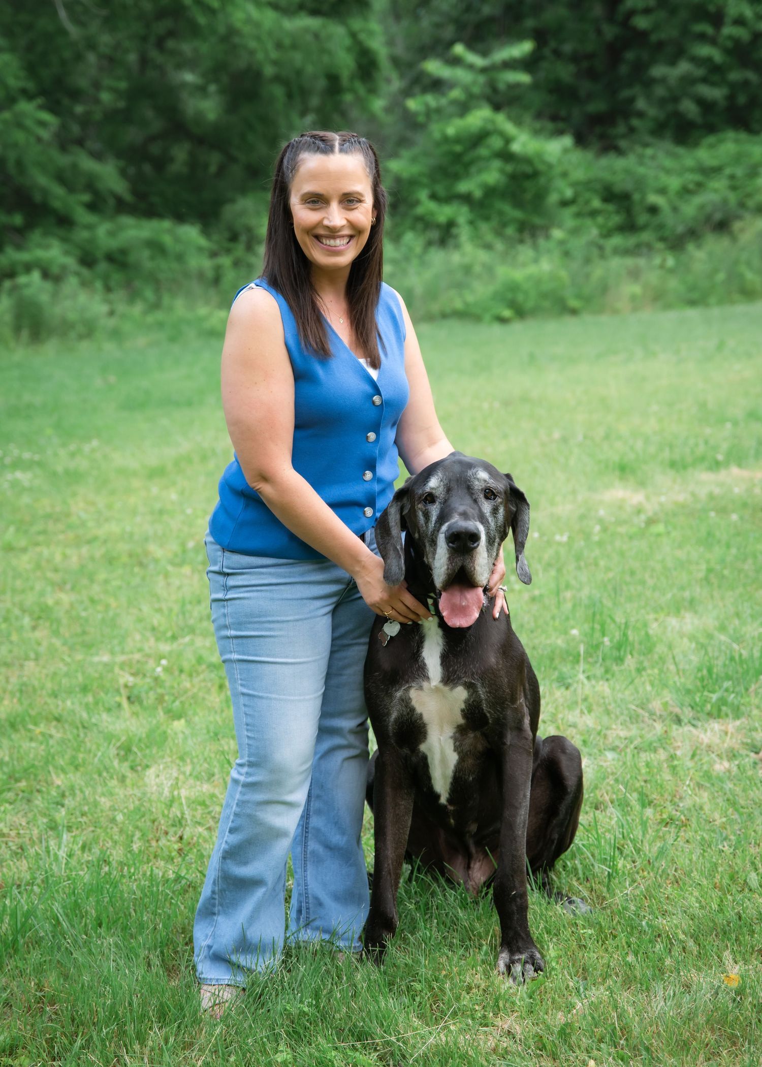 Woman in blue top and jeans smiling beside a large black Great Dane dog on a green grass lawn.