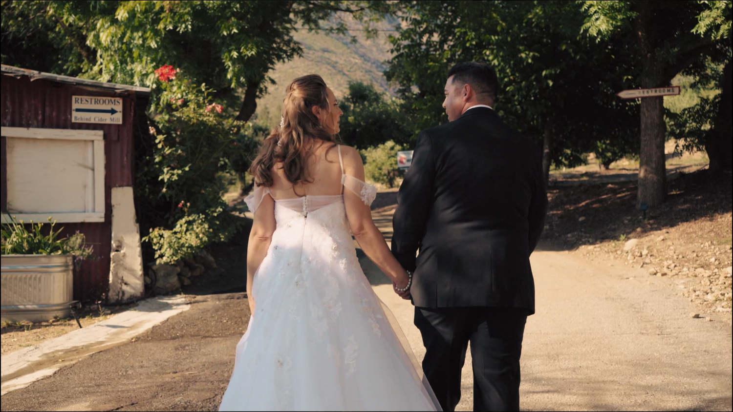 A couple wearing formal wedding attire walk together on a shaded garden path during golden hour.