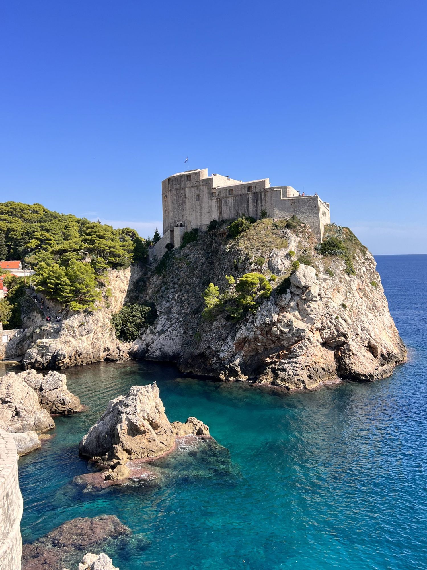 Fort Lovrijenac perched on rocky cliffs above crystal-clear turquoise waters in Dubrovnik, Croatia under a clear blue sky.